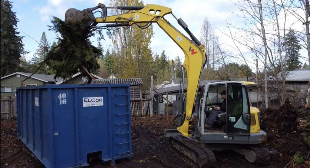 Excavator lifting tree debris into a blue dumpster in a residential area.