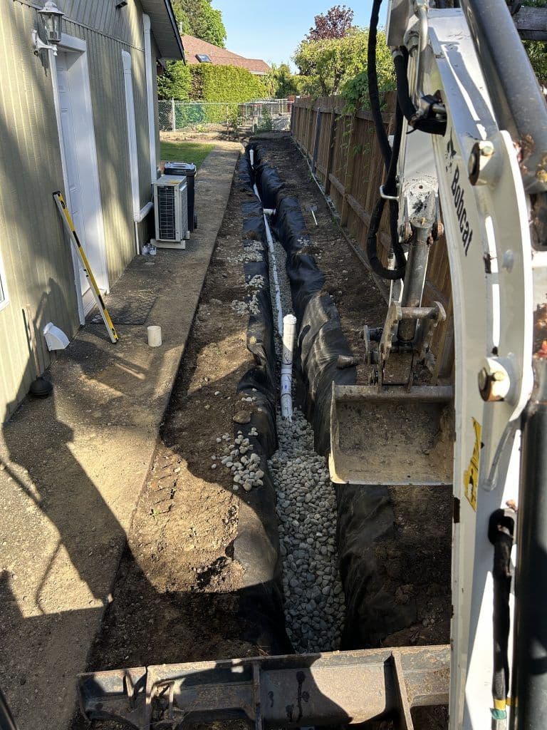 Excavation site showing a trench with gravel, drainage pipe, and construction equipment.