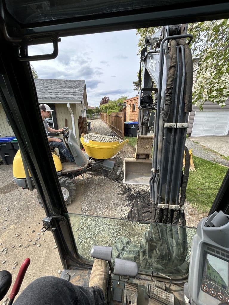 Construction worker operating a mini excavator to load gravel into a yellow wheelbarrow.