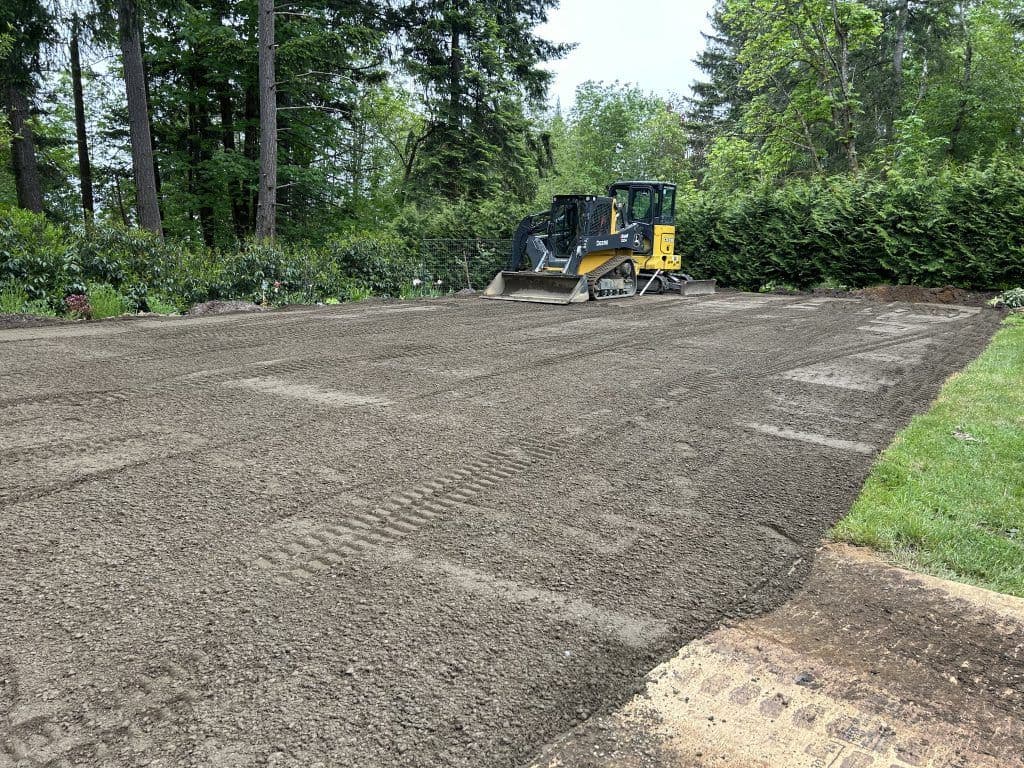 Excavator leveling soil in a landscaped area surrounded by trees and greenery.