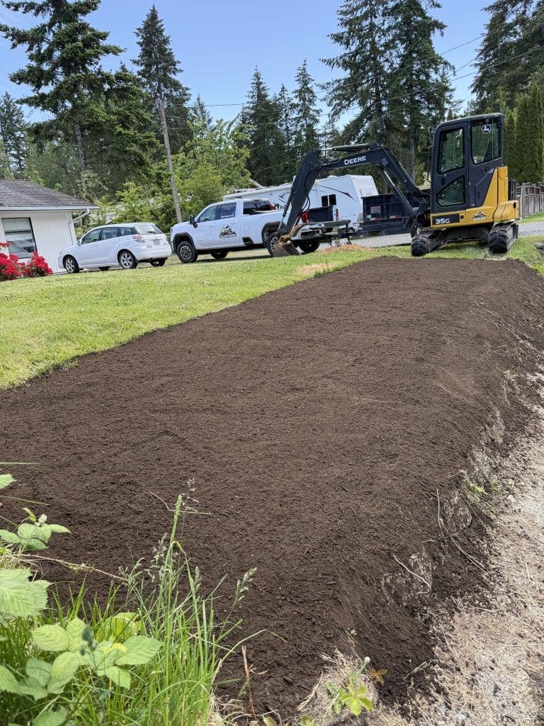 Excavator on freshly tilled soil in a residential yard with parked vehicles and trees.