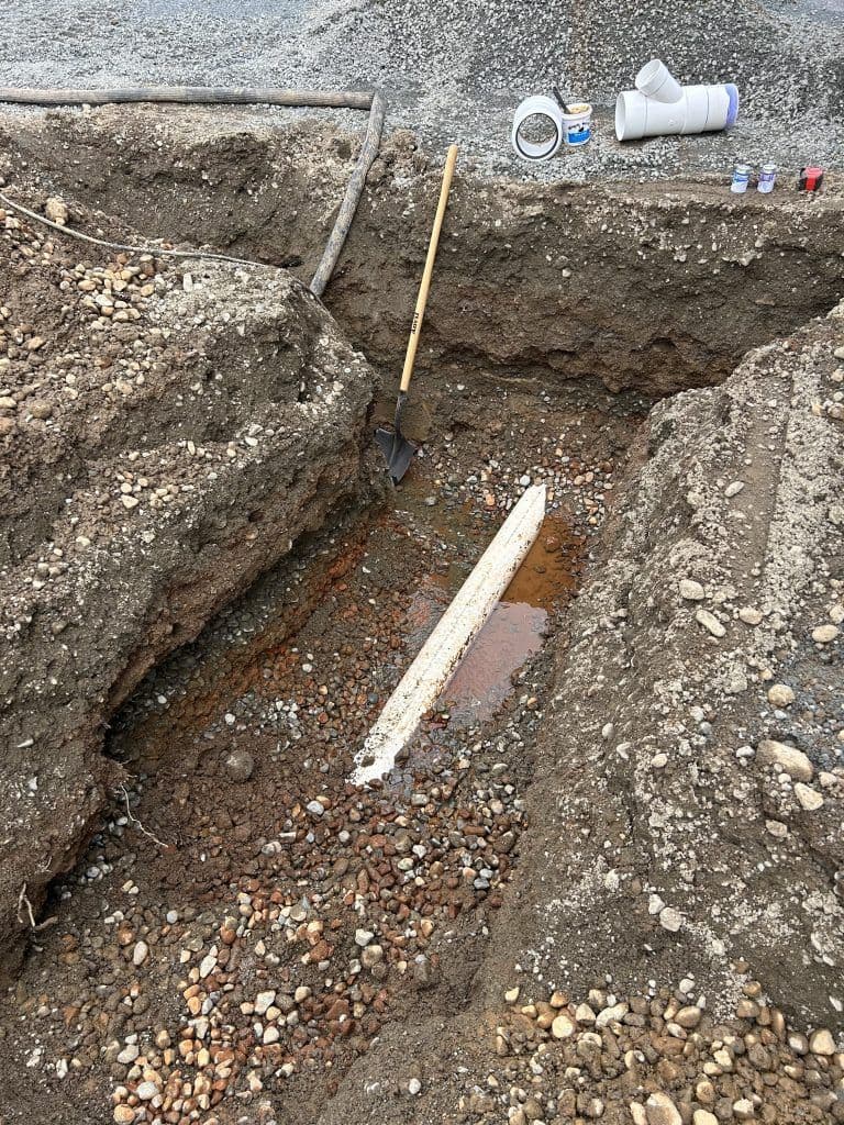 Excavation site with a shovel, drainage pipe, and water pooling in rocky soil.