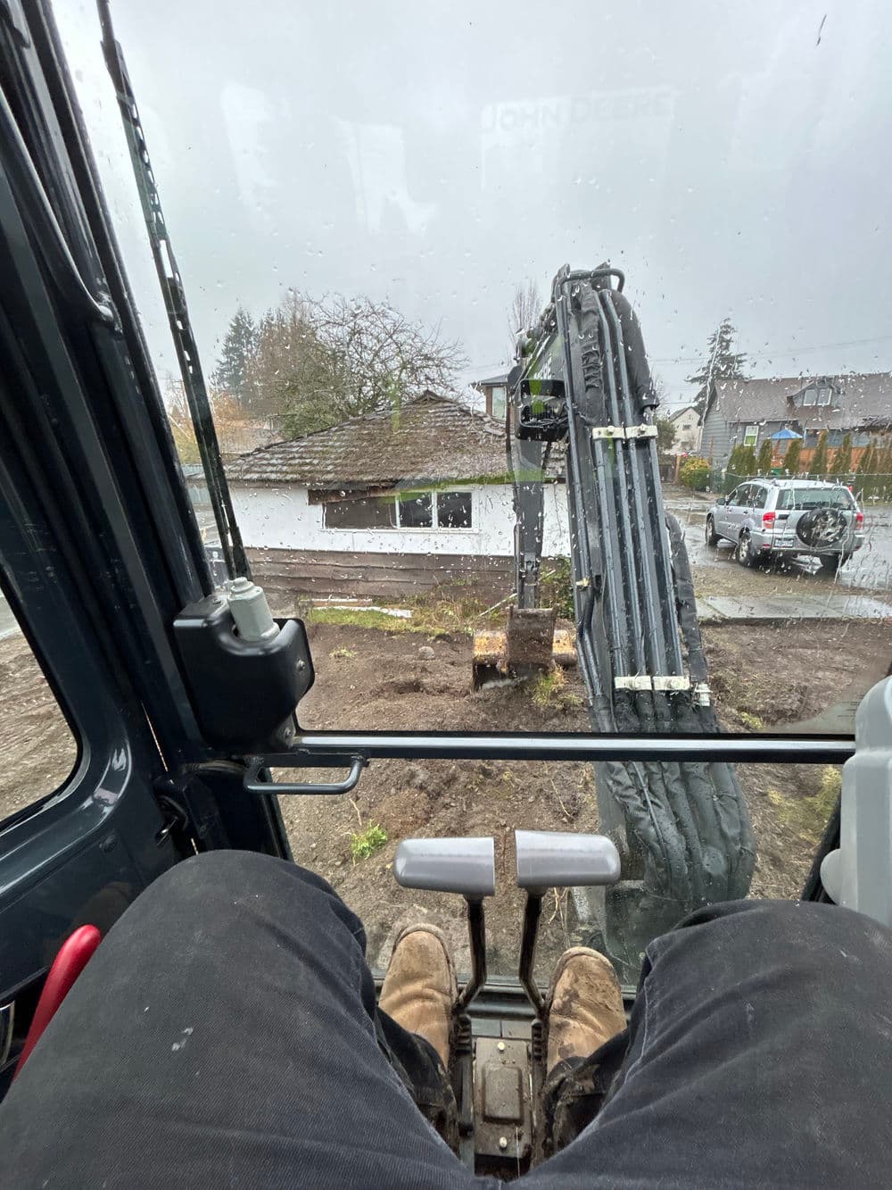 View from inside a John Deere excavator with arm positioned for digging at a construction site.
