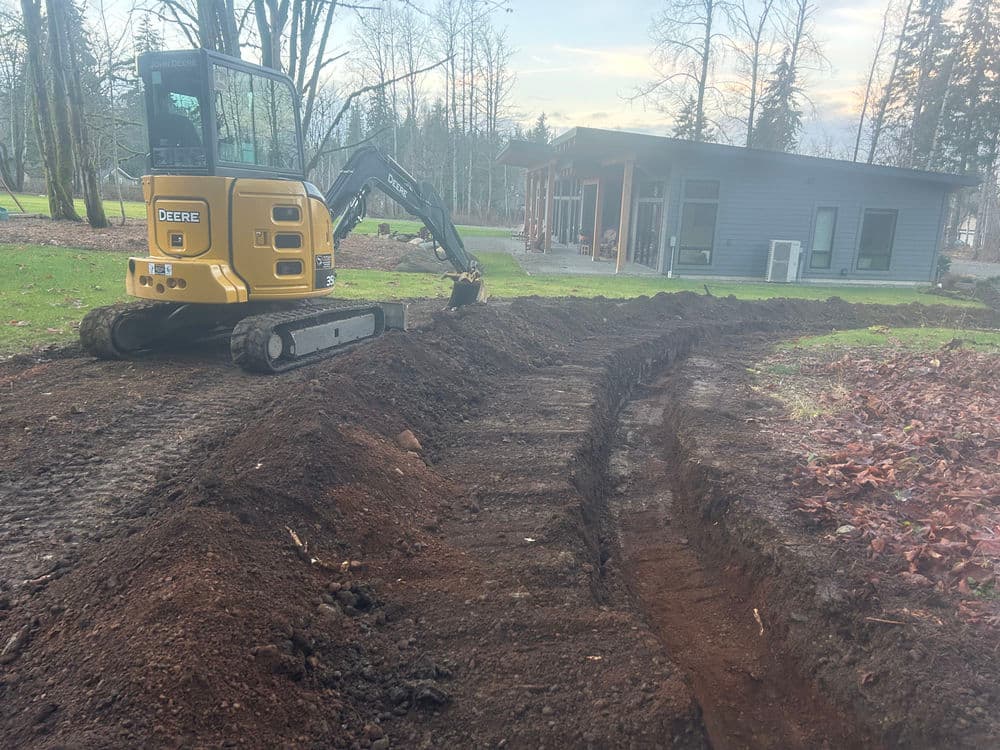 Excavator digging a trench in a yard near a modern house surrounded by trees.