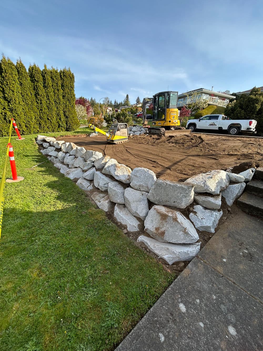 Excavator working on a stone retaining wall in a landscaping project with a grassy area.