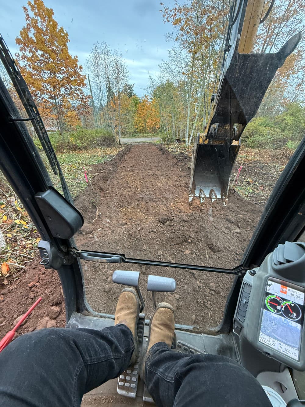 Excavator view showing operator's feet and a cleared path lined with autumn trees.