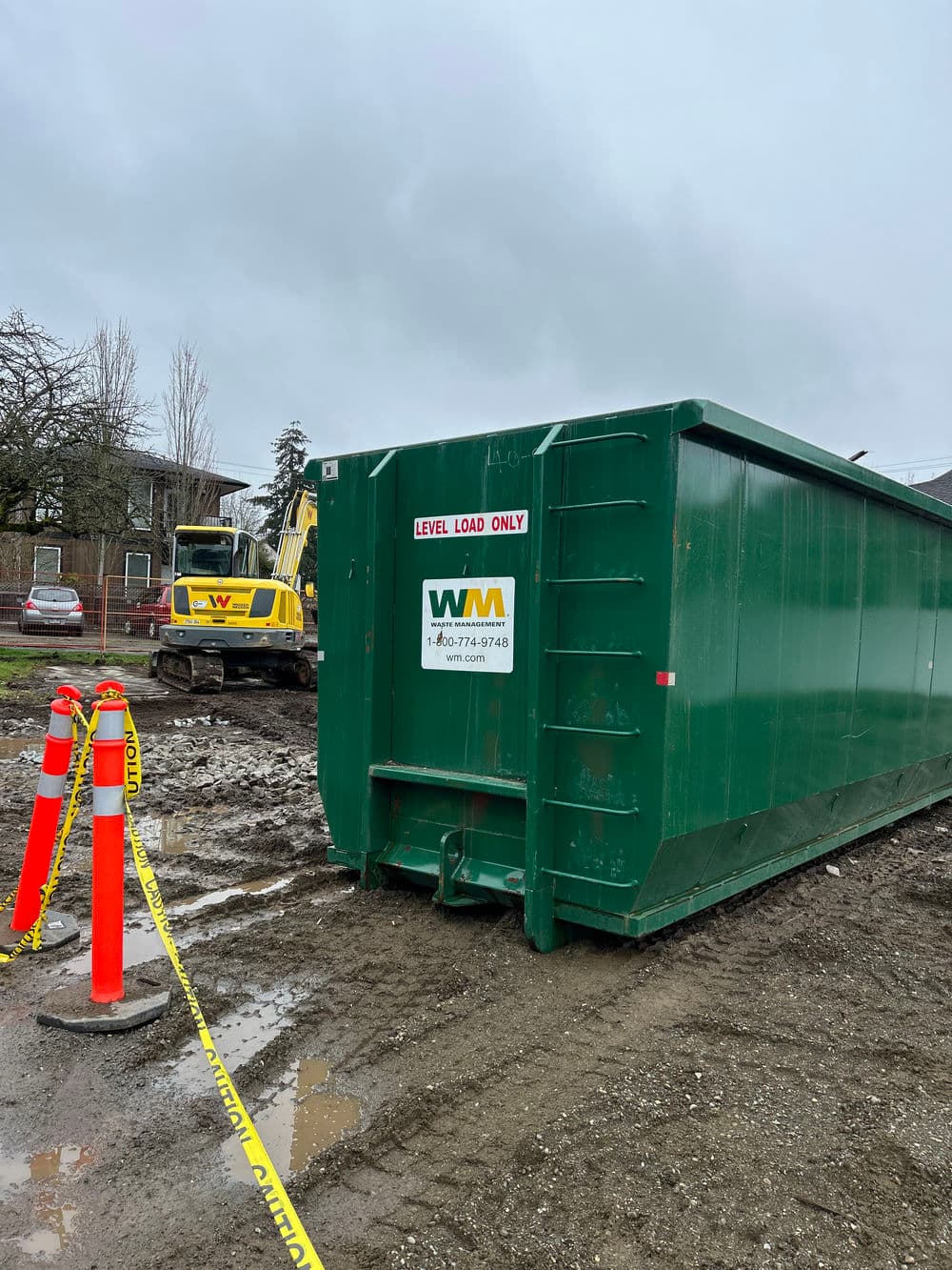 Green dumpster with WM logo on construction site, surrounded by cones and heavy machinery.