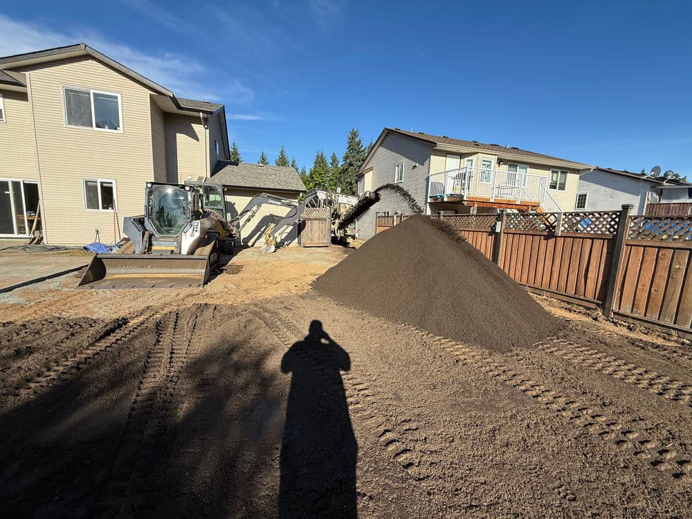 Construction site with earthmoving equipment and a pile of dirt behind residential homes.
