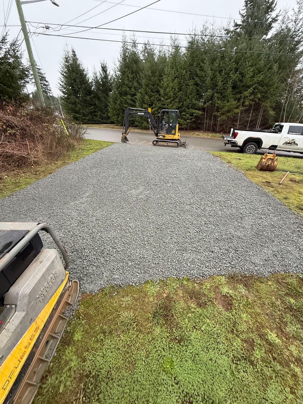 Gravel driveway installation with excavator and truck in forested area.