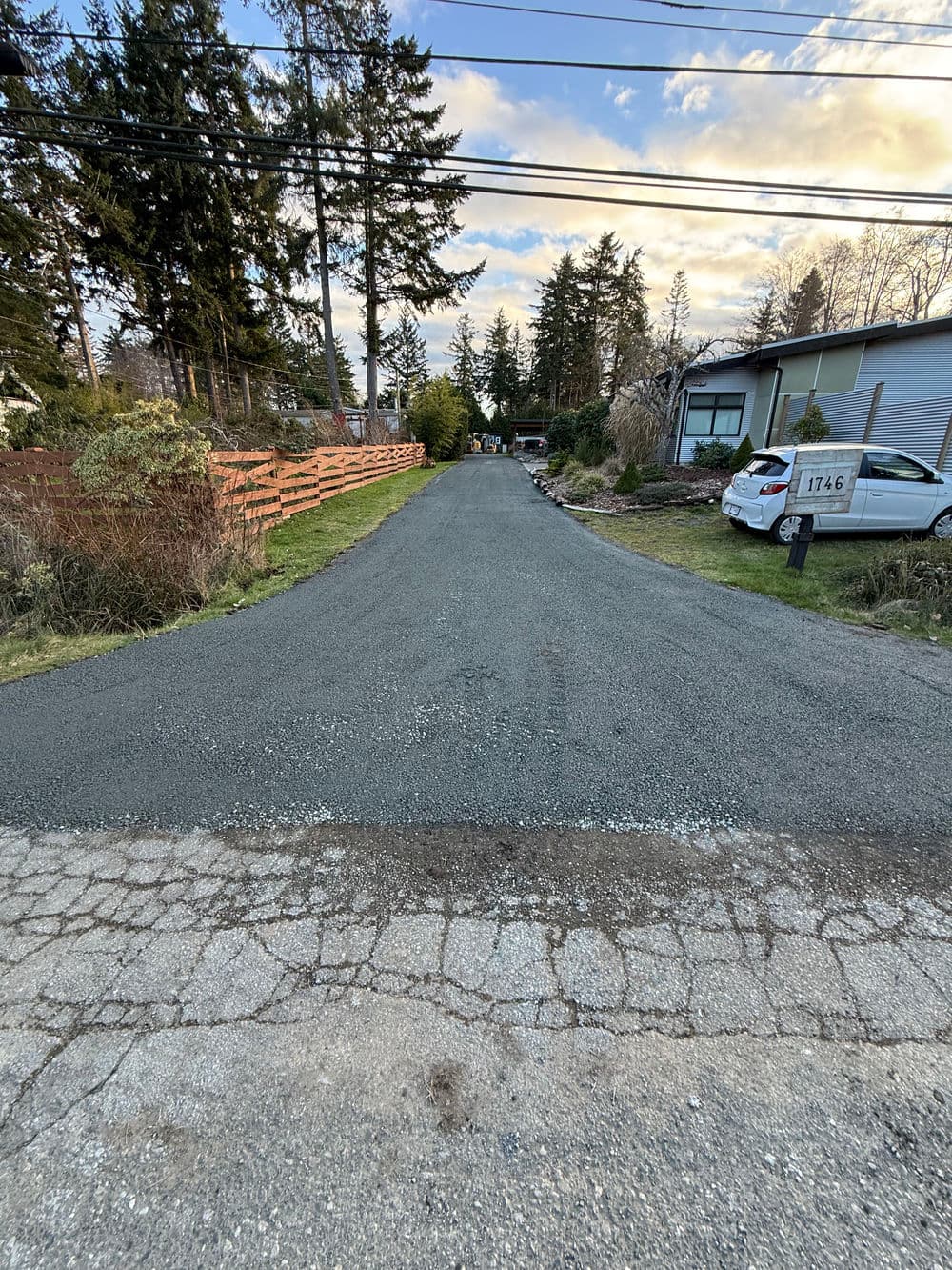 Gravel residential driveway lined with trees and a house at the end, near parked cars.