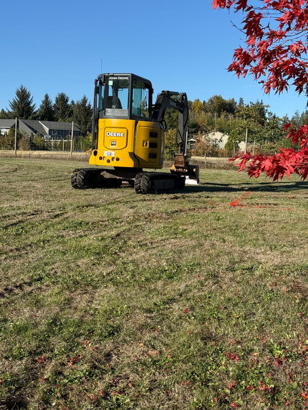 Yellow John Deere excavator on a grassy field with autumn trees in the background.