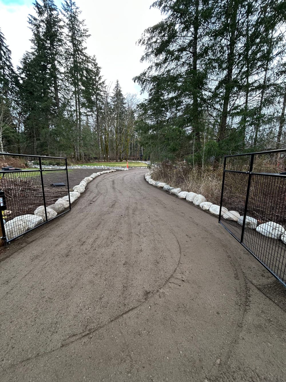 Gravel path through a forested area, lined with rocks and a metal fence on both sides.