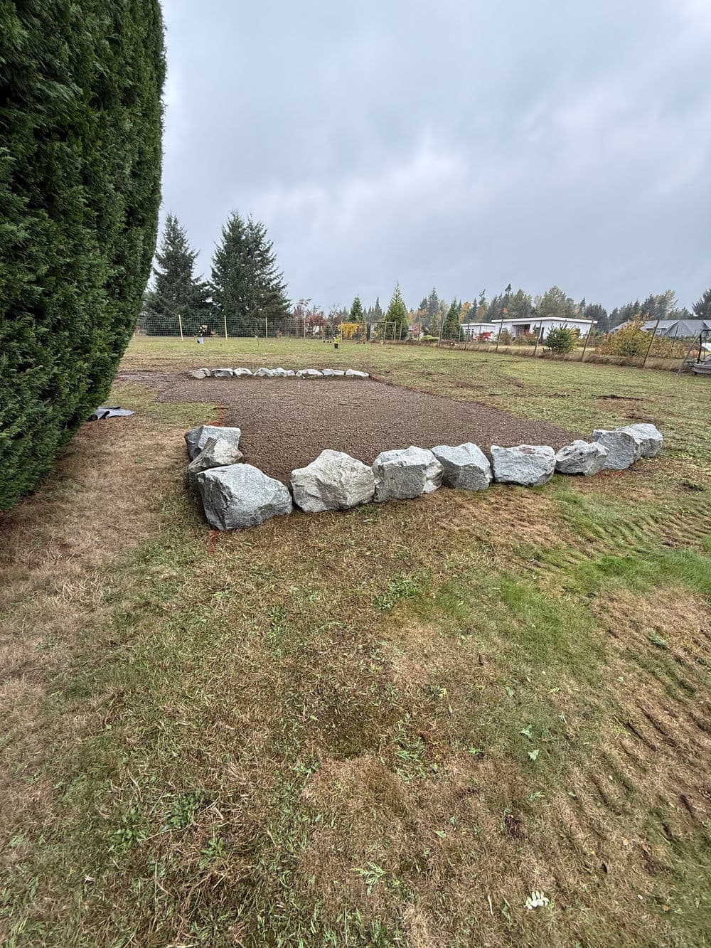 Cleared garden area enclosed by large rocks, surrounded by grass and trees on an overcast day.