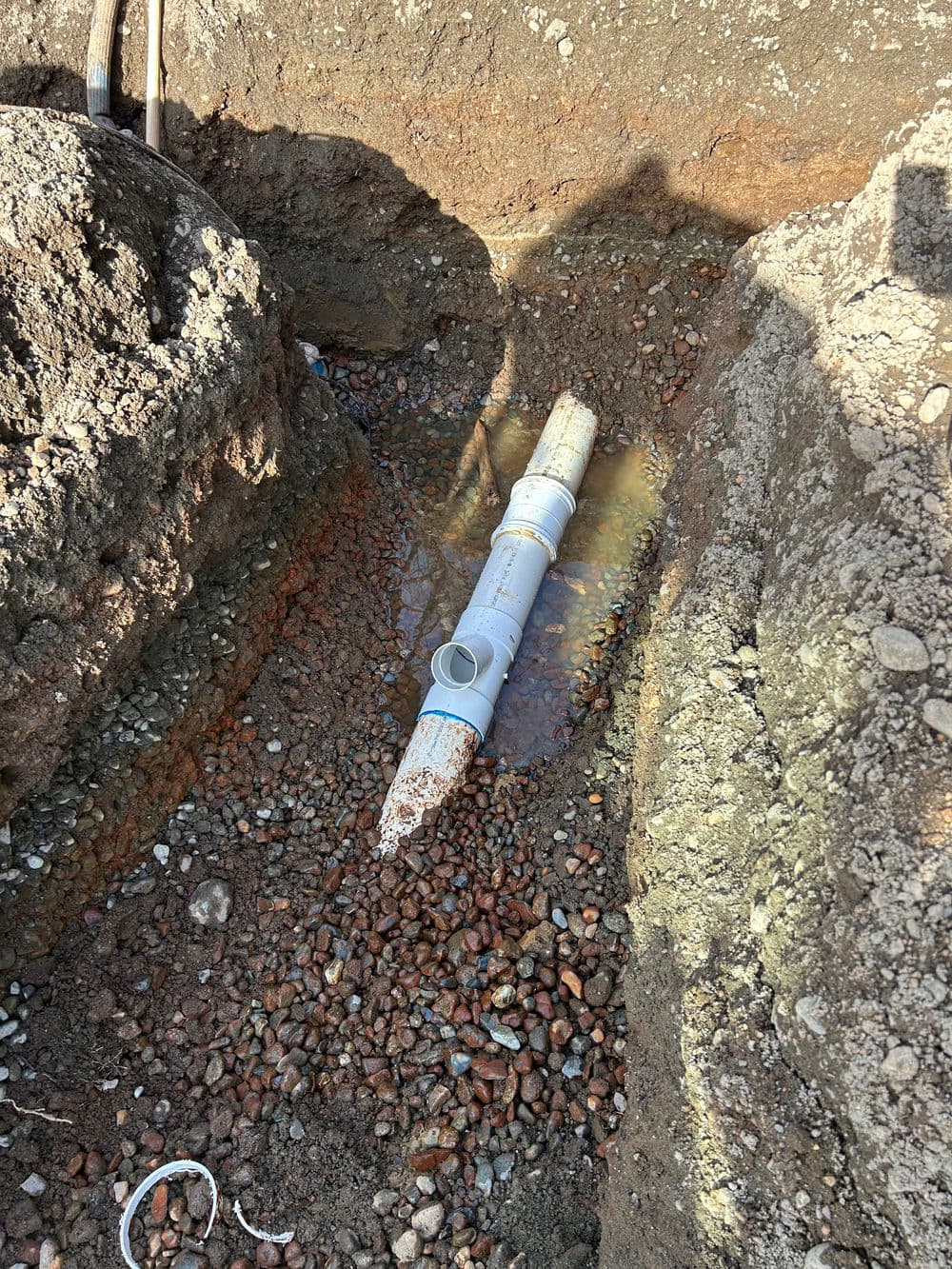 Broken PVC pipe in a trench with standing water and gravel around it. Garden repair project.