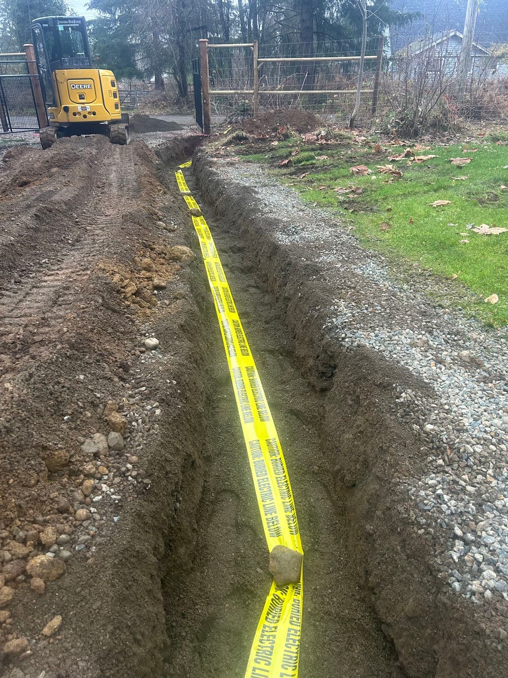 Excavation trench marked with caution tape, heavy machinery nearby, in a rural setting.