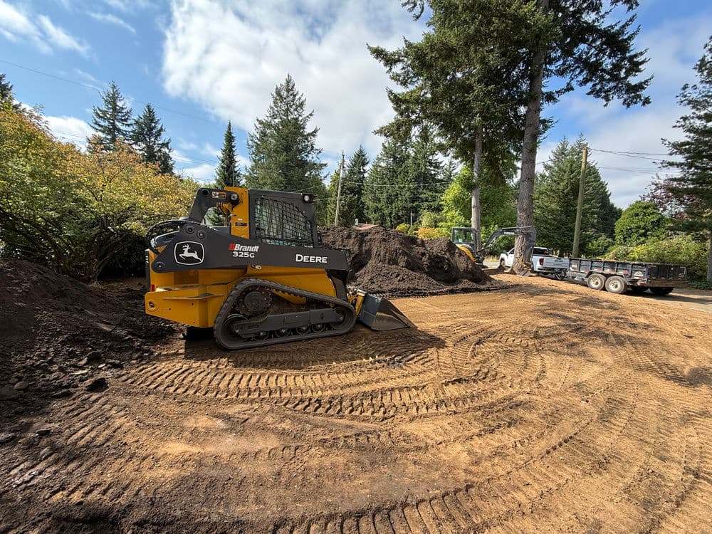 John Deere 325G compact track loader on a construction site with trees and soil piles.