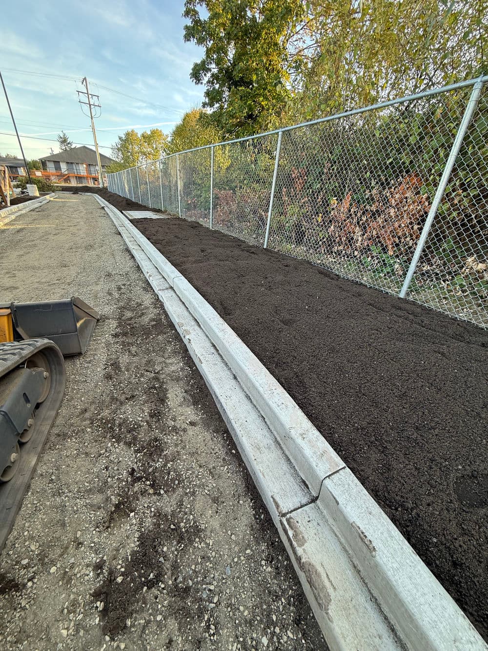 Newly paved sidewalk with dark soil alongside a chain-link fence and trees in the background.