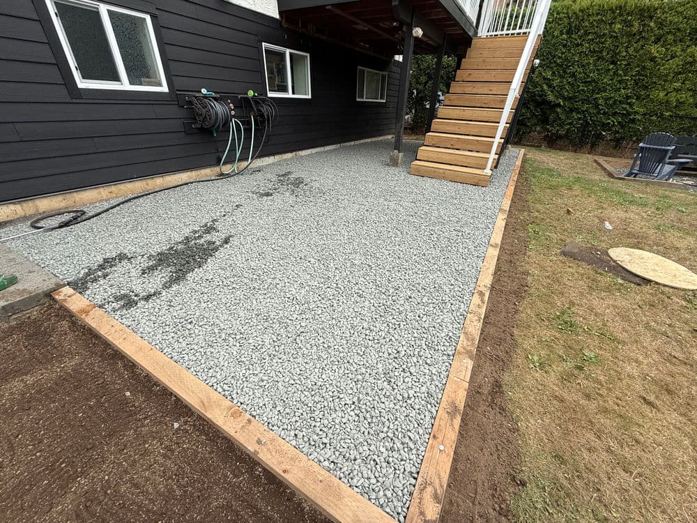 Gravel patio area under a house with wooden steps, hoses, and landscaped surroundings.