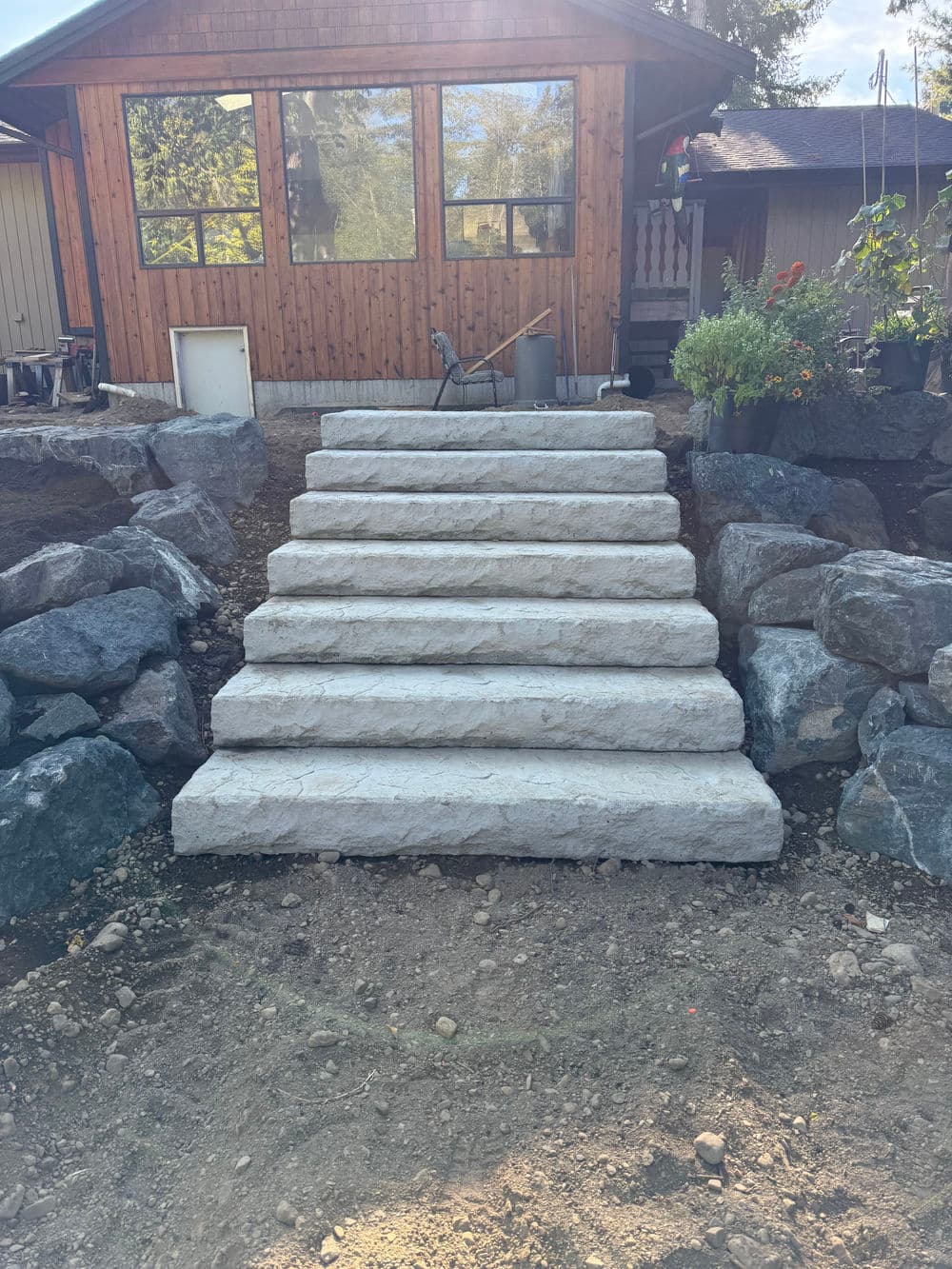 Concrete steps leading up to a wooden house, surrounded by landscaped rocks and plants.
