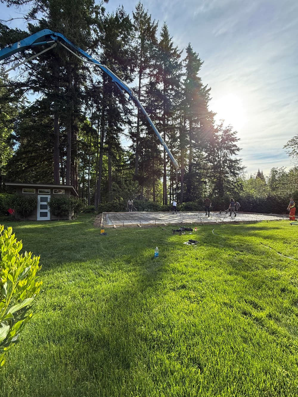 Construction crew pouring concrete in grassy area surrounded by tall trees and sunlight.
