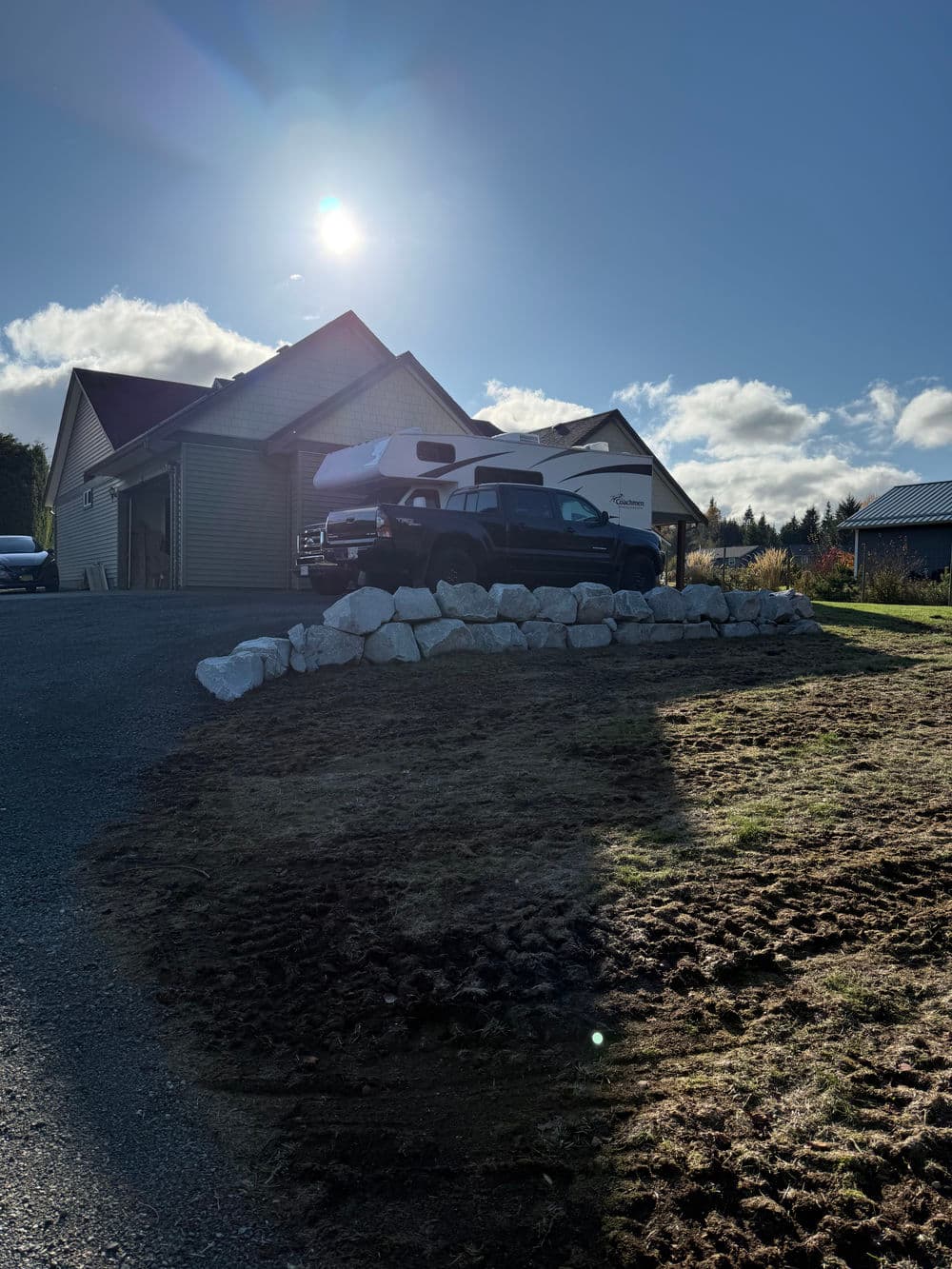 Pickup truck parked near a house under a sunny sky, with a stone wall in the foreground.