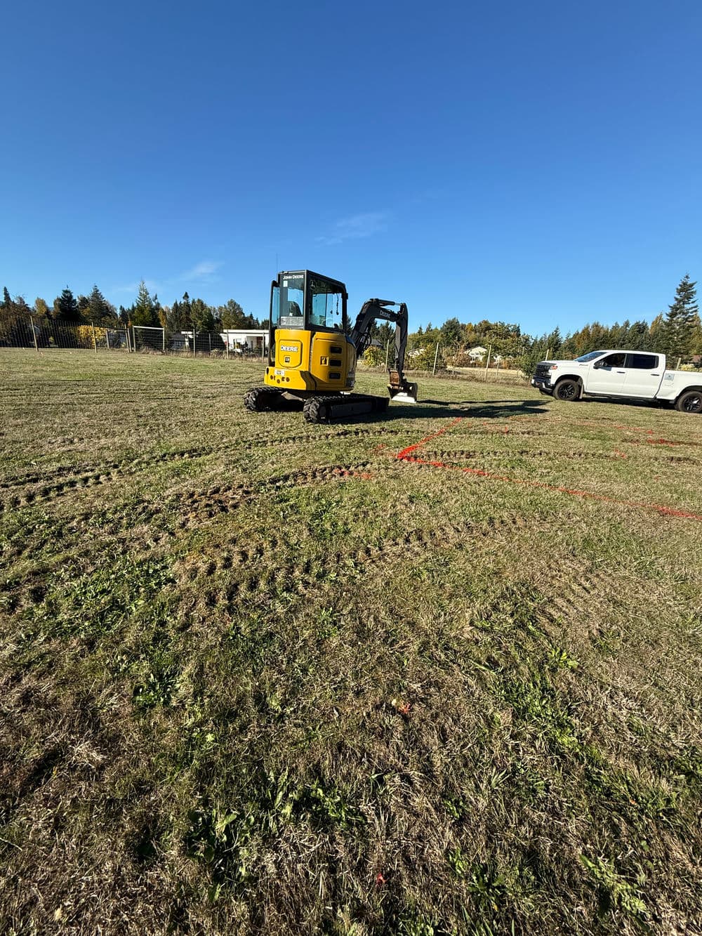 Excavator working on a grassy field with tire tracks and clear blue sky.