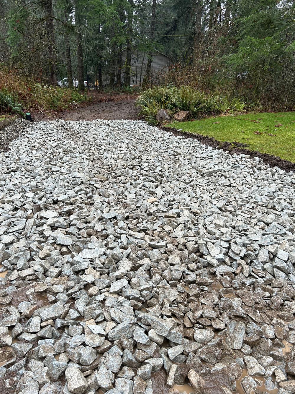 Gravel pathway under construction in a wooded area, surrounded by greenery and trees.