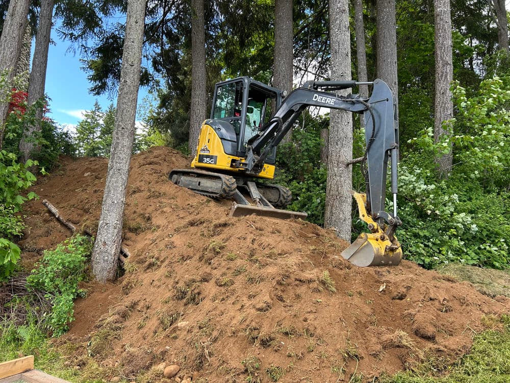 Excavator on hillside surrounded by trees, clearing soil for construction project.
