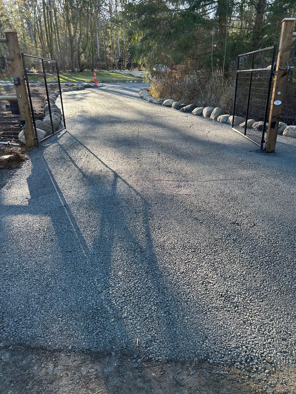 Gravel driveway entrance with wooden gates and shadows on a sunny day amidst greenery.