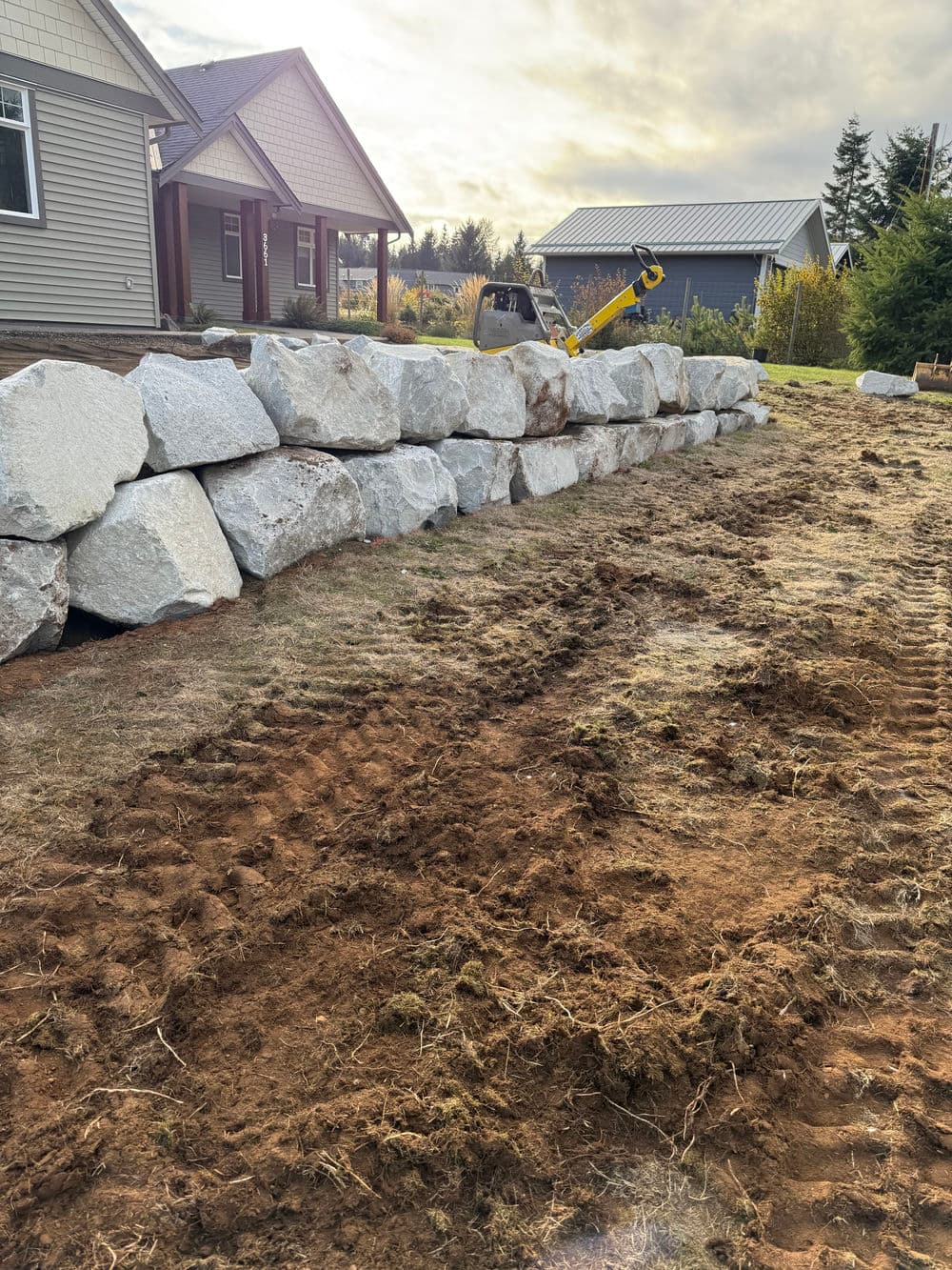 Stone wall construction in a residential yard with freshly turned soil and a home in the background.