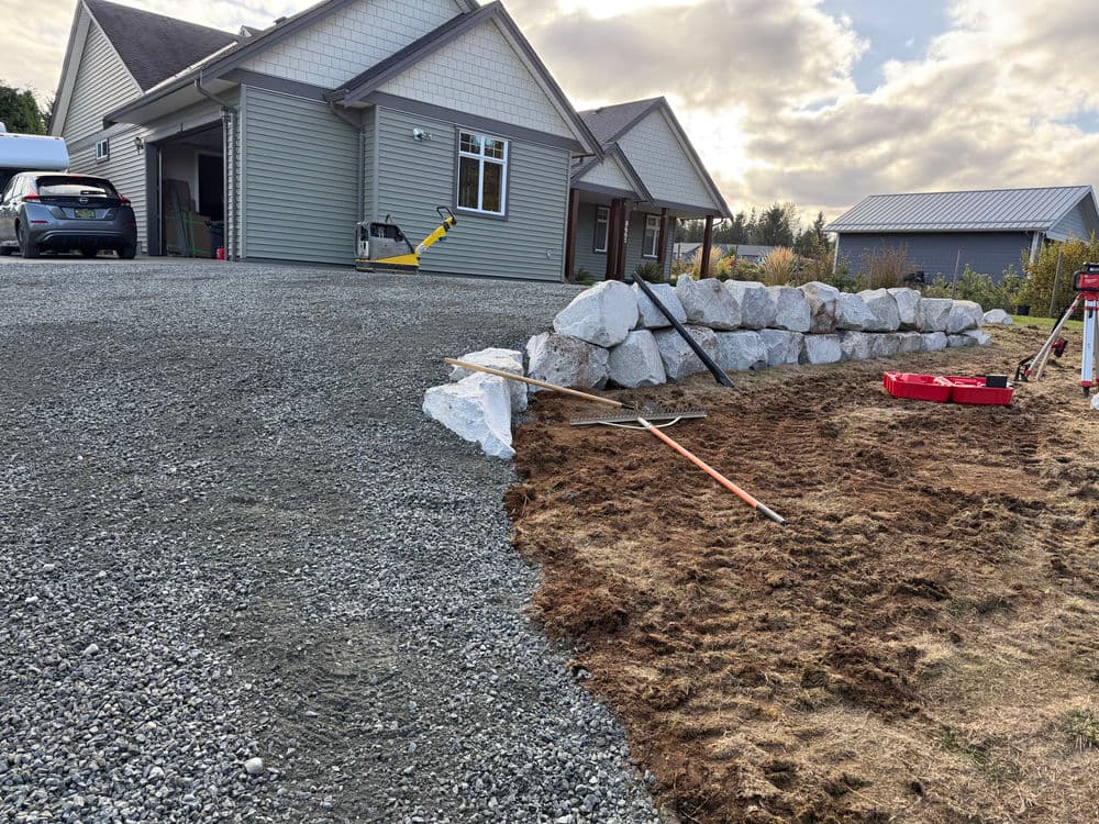Residential landscape featuring gravel driveway, rock wall, and new home under construction.