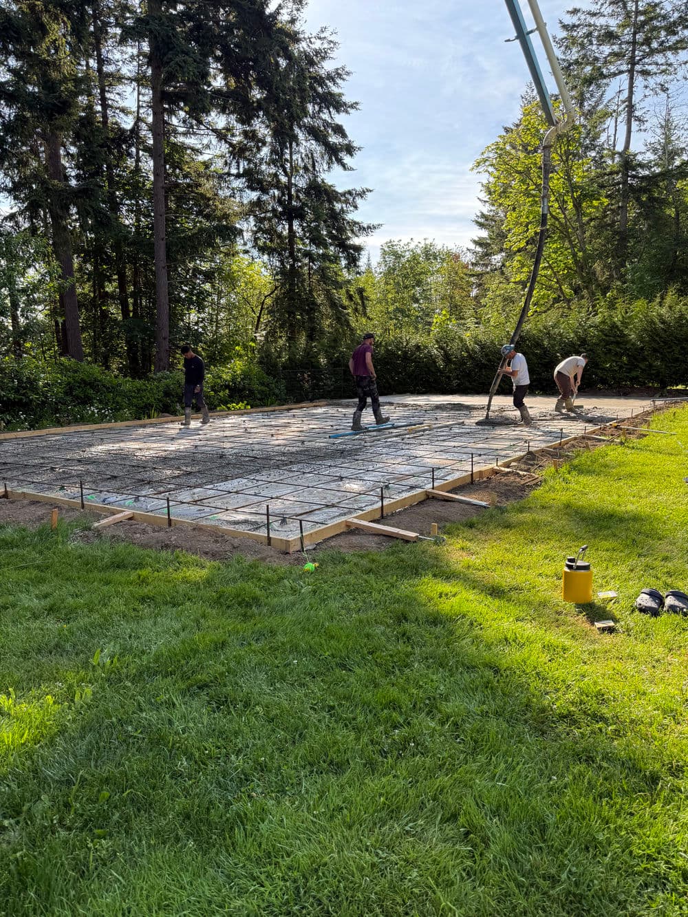 Workers pouring concrete for a foundation in a wooded area on a sunny day.