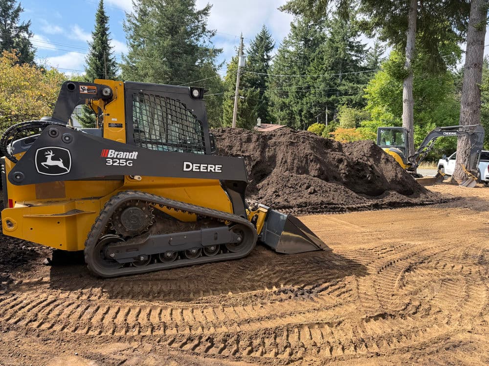 John Deere 325G skid steer loader working on a construction site with soil piles.
