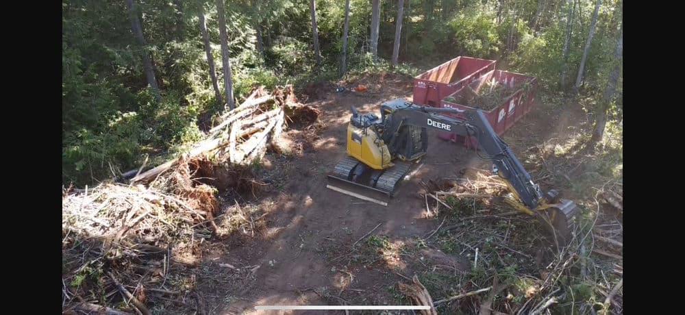 Excavator clearing a forested area with a debris container in a construction site.
