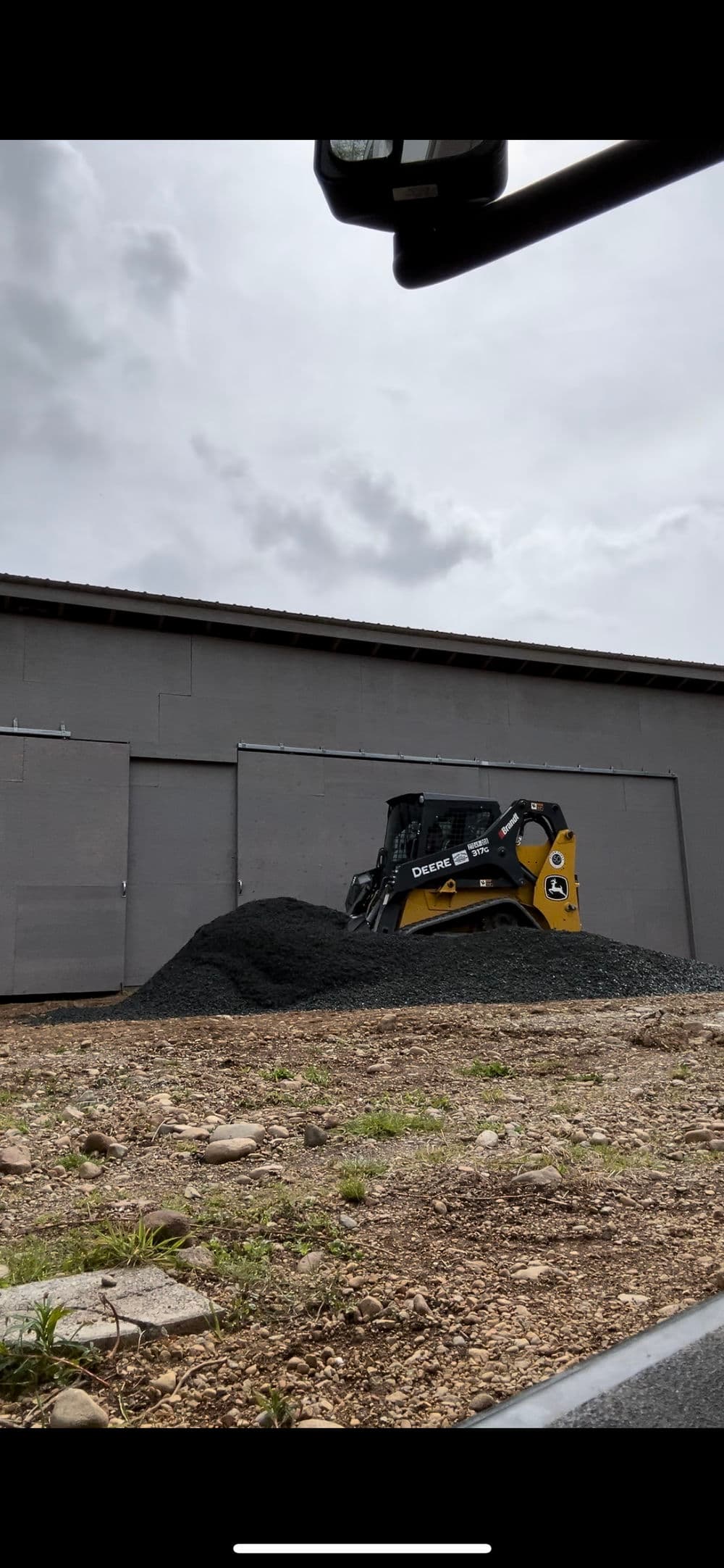 Skid steer loader on a gravel mound beside a gray building under cloudy skies.